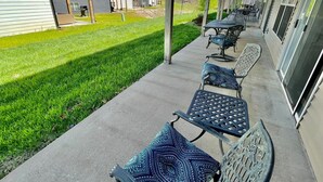 Un espace extérieur aménagé avec des sièges, protégé par un toit, comprenant des chaises en métal et des coussins à motifs, situé à côté d’un bâtiment doté de grandes fenêtres et offrant une vue sur une pelouse verdoyante et d’autres bâtiments.