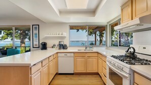 Una cocina moderna con lavavajillas, estufa y lavabo, ventanas amplias con vista al océano, y armarios de madera.