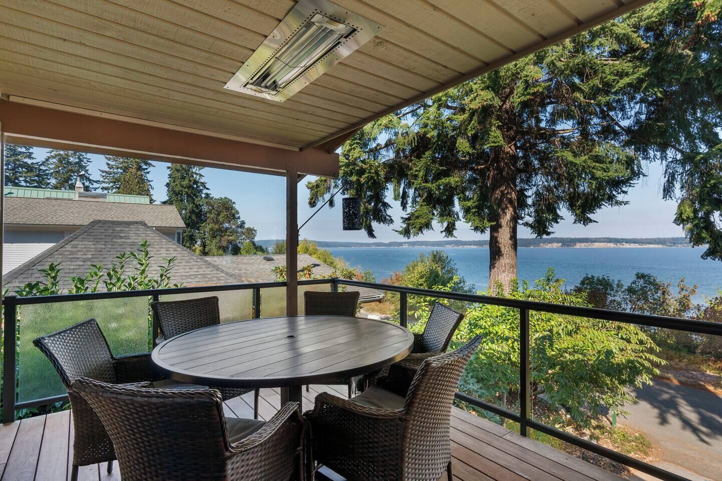A covered outdoor seating area with a table and chairs, overlooking a body of water and a forested area.