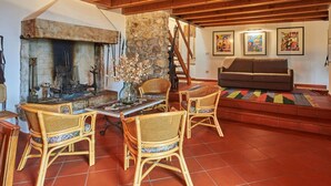 A cozy living room with a stone fireplace, wooden beams, wicker furniture, and colorful artwork.