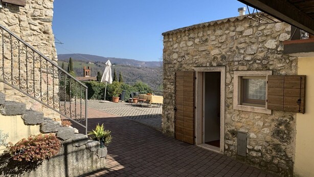 A stone building with a wooden door, a small window, and a paved patio area with outdoor furniture.
