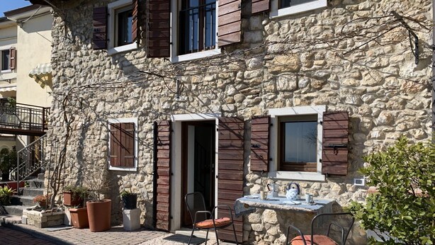A stone building with wooden shutters, a small outdoor seating area with a table and chairs, and potted plants.
