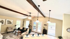 A spacious living room with a high ceiling, a ceiling fan, and pendant lights. There is a large window with blinds, a fireplace, and a kitchen area with a bar counter.