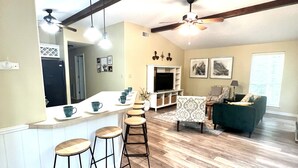 A modern kitchen with wooden bar stools, a dining area with a patterned table, and a living room with a green sofa.