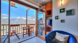 A balcony with a wooden table and chairs, a blue sofa with a decorative pillow, and a view of mountains through large glass doors. - Rebecca's House among the greenery, Bardonecchia, Italy (Bardonecchia)