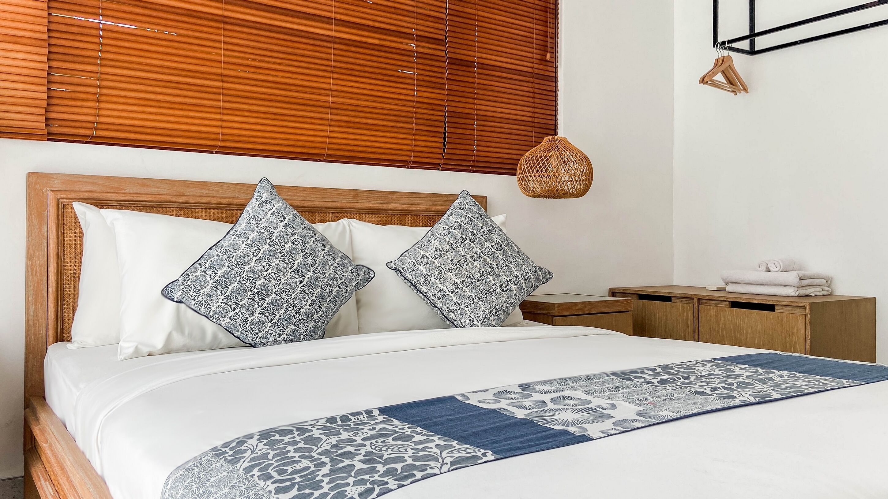 A neatly made bed with patterned pillows, a wooden headboard, a bedside table with a woven lamp, and a wooden nightstand.