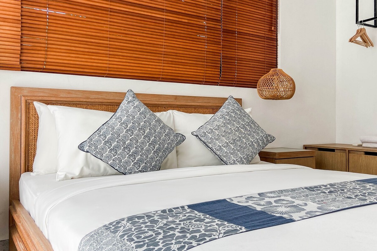 A neatly made bed with patterned pillows, a wooden headboard, a bedside table with a woven lamp, and a wooden nightstand.