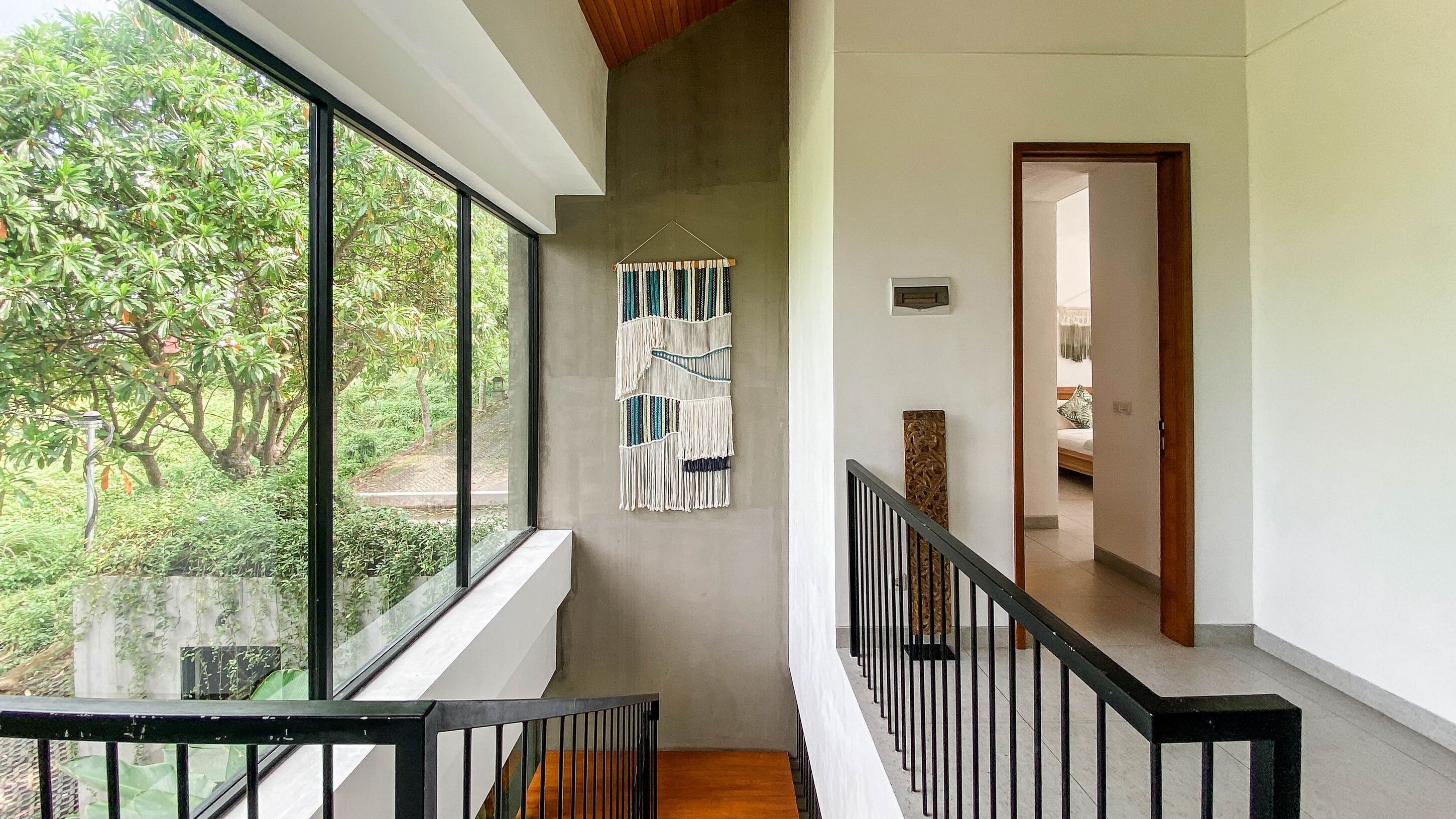 A modern interior with a staircase leading to a bedroom. A macrame wall hanging is visible, and a view of greenery is seen through large windows.