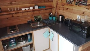 Une cuisine compacte aux murs en bois, équipée d’un évier, d’une cuisinière, d’un four à micro-ondes et d’une étagère où sont rangées des tasses et des bols.