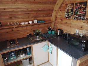A compact kitchen with wooden walls, a sink, a stove, a microwave, and a shelf with cups and bowls.