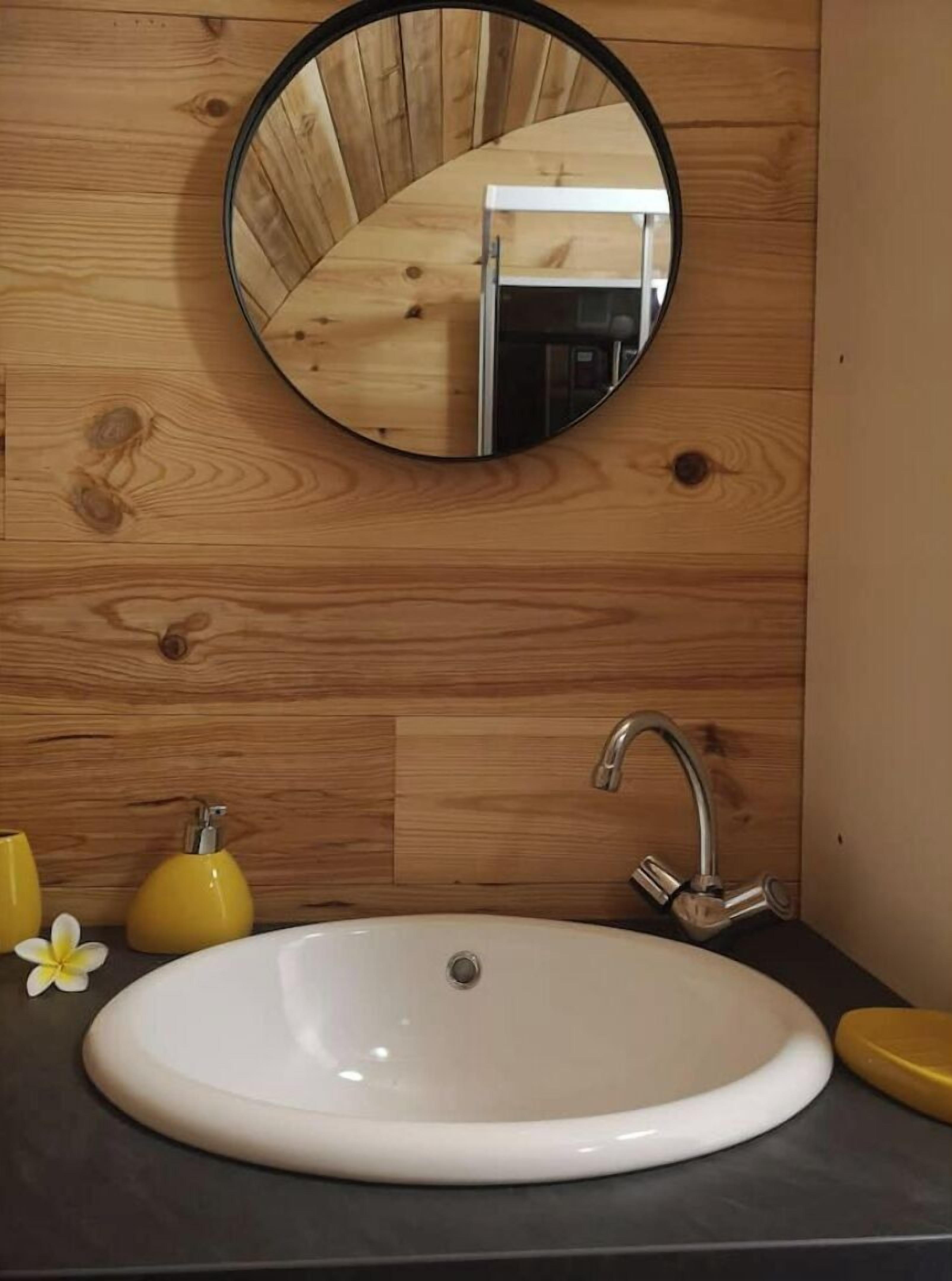 Een badkamer met een houten wand, een ronde spiegel, een witte wastafel, een moderne kraan en gele zeepdispensers.