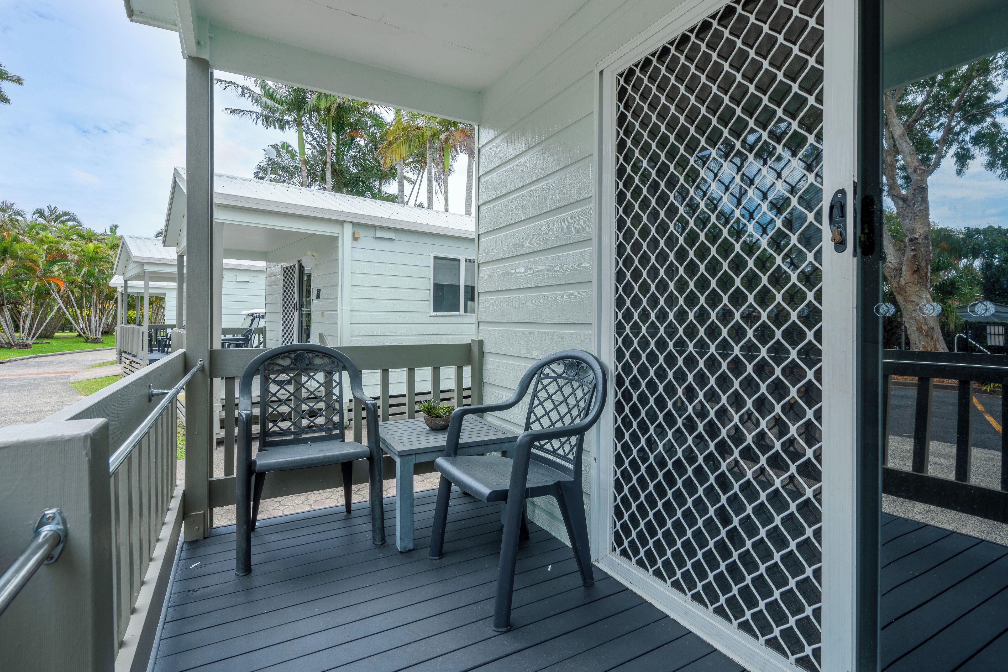 A covered outdoor area with a table and two chairs, a metal fence, and a view of a white building with a window.