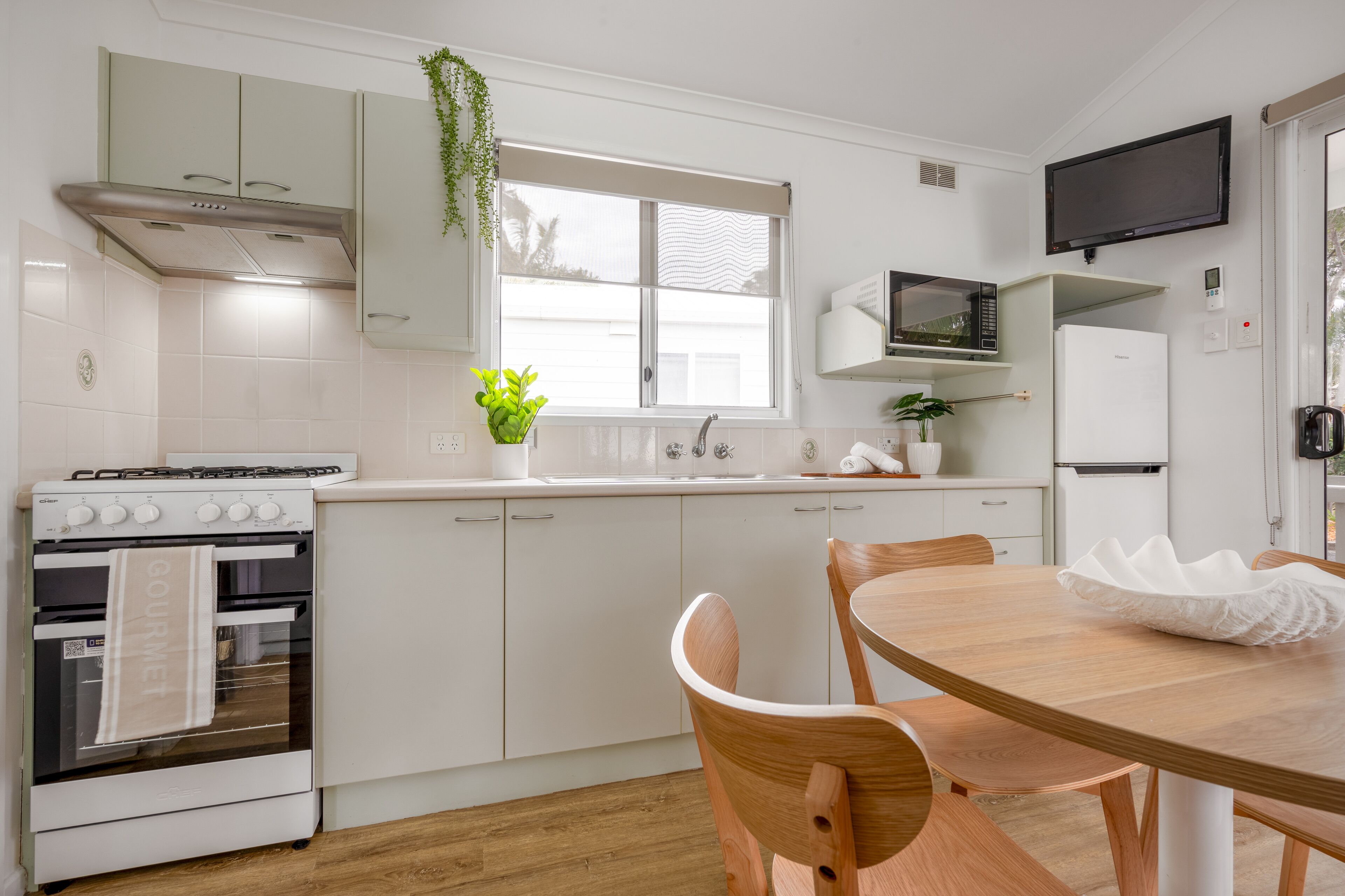 A modern kitchen with white cabinetry, a stove, a microwave, and a refrigerator. There is a dining table with chairs and a potted plant on the windowsill.