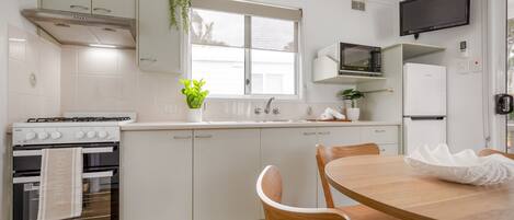 A modern kitchen with white cabinetry, a stove, a microwave, and a refrigerator. There is a dining table with chairs and a potted plant on the windowsill.