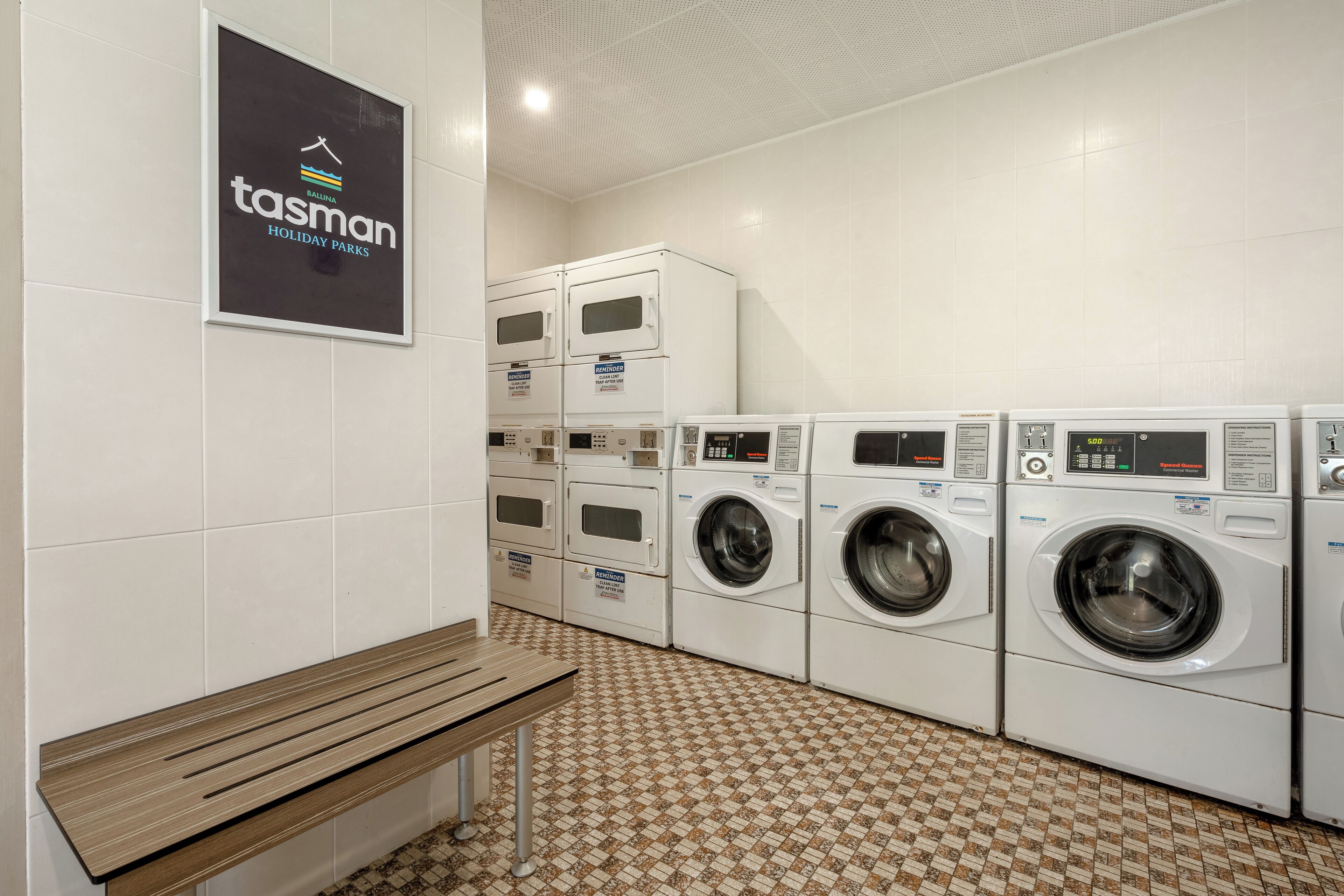 A laundry facility with multiple washing machines and dryers, a bench, and a sign for Tasman Holiday Parks.