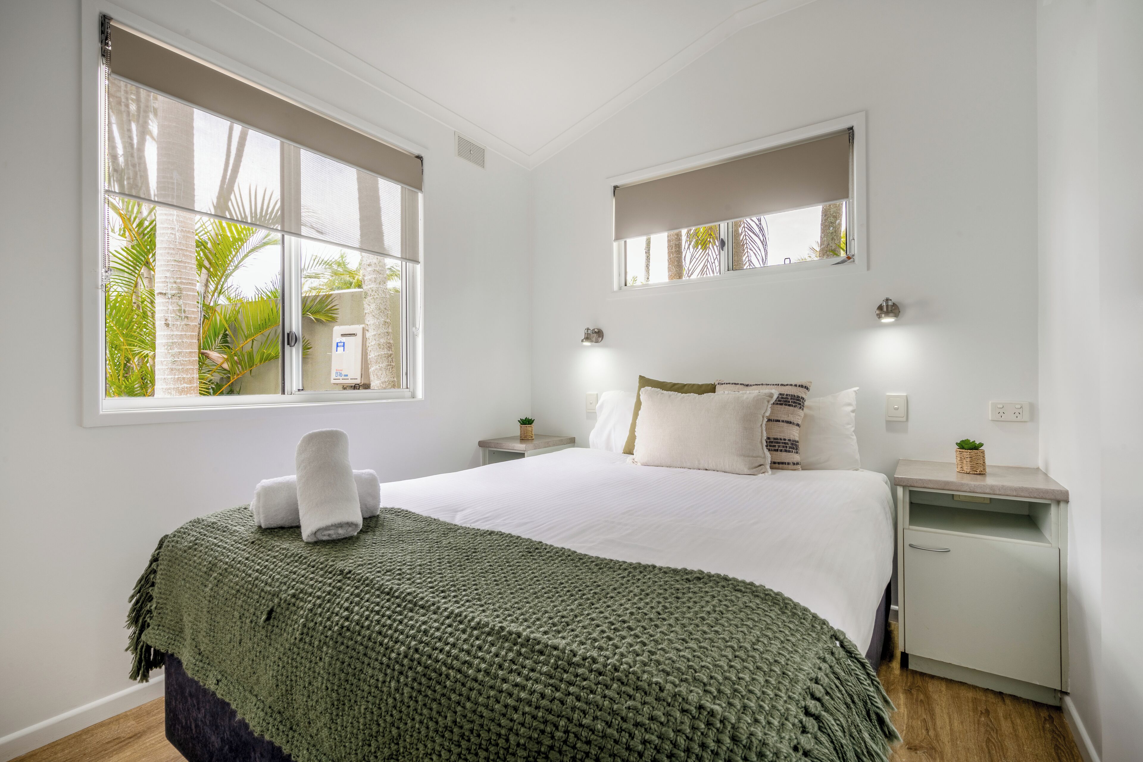 Ein ordentlich eingerichtetes Schlafzimmer mit einem großen Bett, einem Nachttisch, einer kleinen Kommode und einem Fenster mit Blick auf Grünflächen.