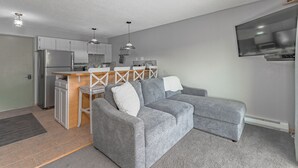 Een ruimtelijk ingerichte leefruimte met een kitchenette, een grijze bank en een aan de muur bevestigde televisie.