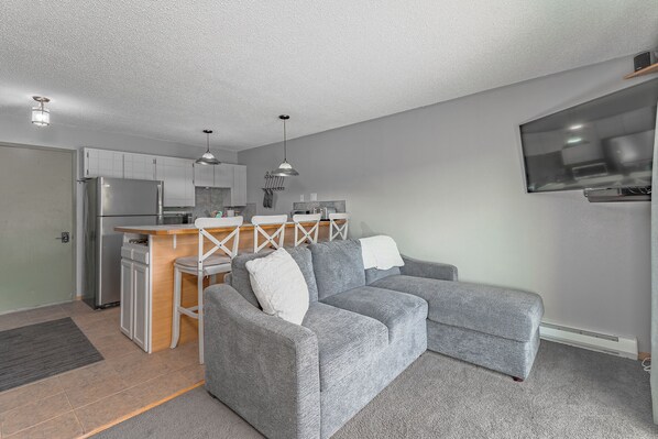 A compact living space with a kitchenette, grey sofa, and a TV mounted on the wall. - Schweitzer Mountain View Condo (Sandpoint)