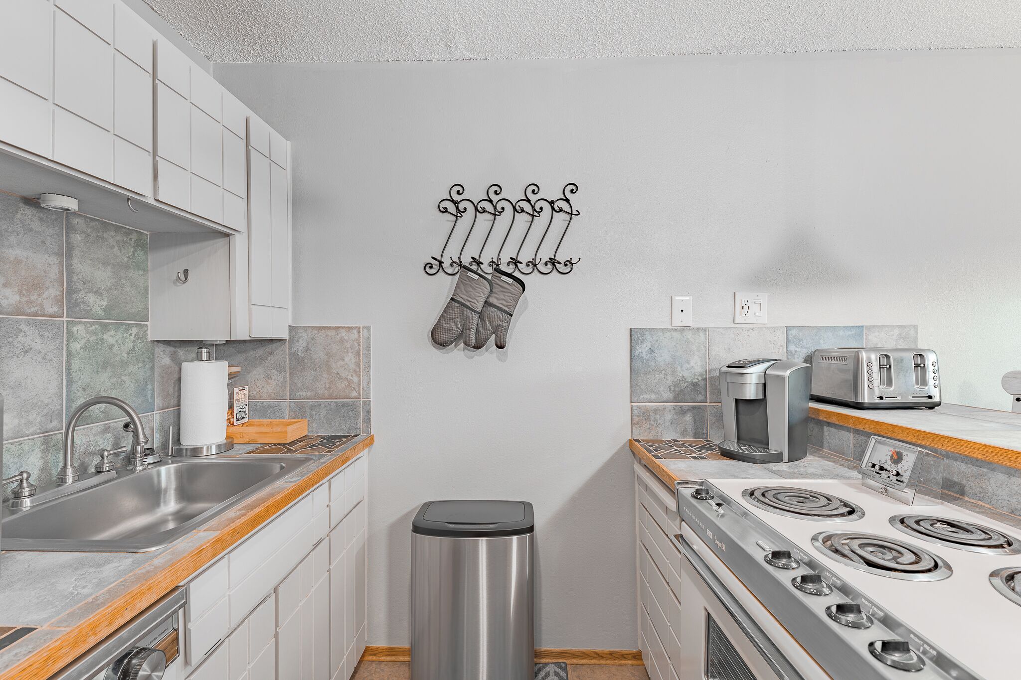 A modern kitchen with white cabinets, a stainless steel sink, a trash bin, a stove, and a toaster.