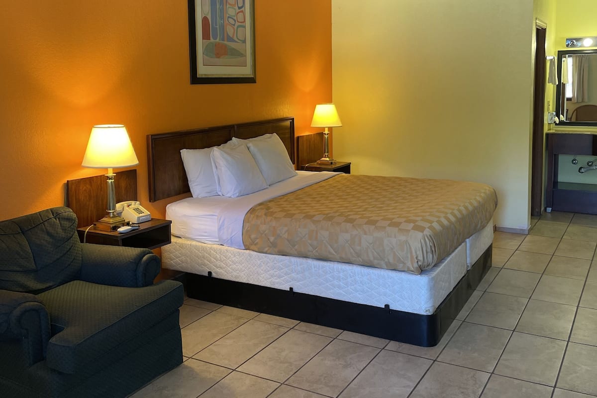 A hotel room with a bed, two bedside lamps, a chair, a nightstand, a painting on the wall, and a bathroom visible through an open door.