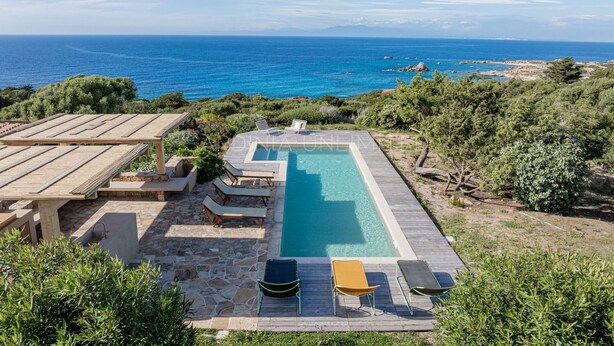 Ein rechteckiger Pool mit Holzterrasse und Liegestühlen, umgeben von Grün und mit Blick auf die Küste.