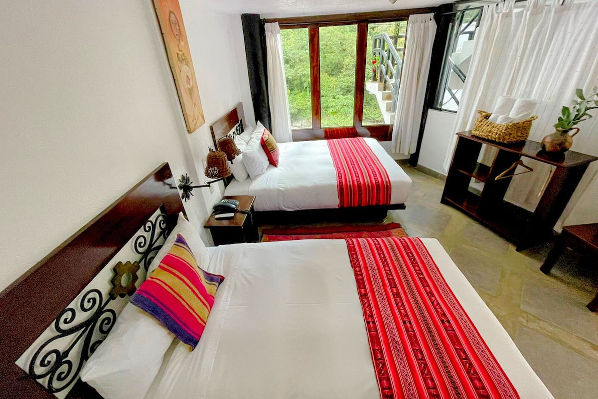 Habitación de hotel con dos camas, una mesita, una silla, un ventanal con cortinas y vistas al exterior con vegetación.