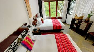 A hotel room with two beds, a small table, a chair, a window with curtains, and a view of greenery outside.