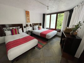 Habitación de hotel con dos camas, un ventanal con cortinas, una mesita con un florero y una obra de arte colgada en la pared.