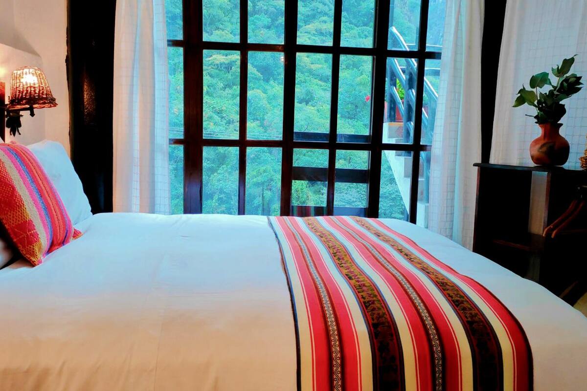Una cama grande con sábana blanca y manta a rayas de colores, una lámpara de noche y vistas de un paisaje verde y exuberante a través de una ventana.