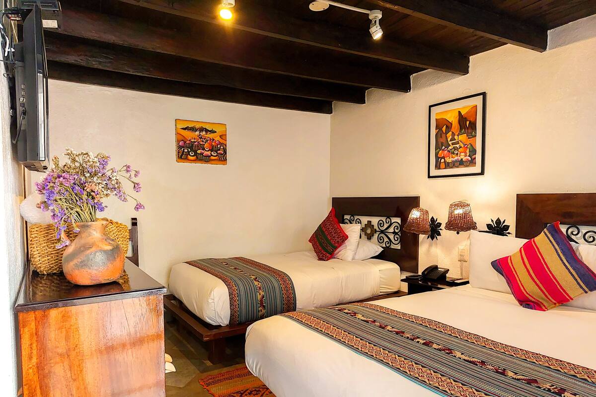 Habitación de hotel con dos camas, una mesita de noche de madera, un jarrón con flores y dos cuadros colgados en la pared.