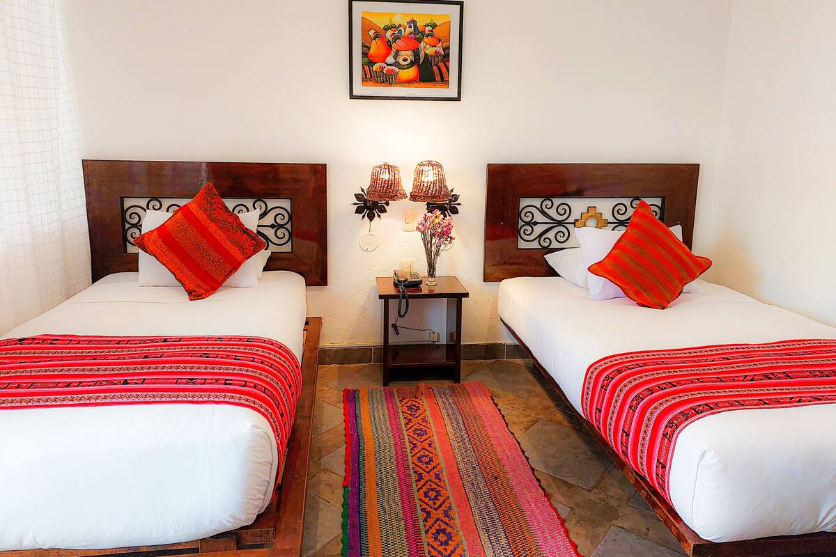 Habitación de hotel con dos camas, una mesita de noche con lámpara, un cuadro enmarcado en la pared y una alfombra estampada.