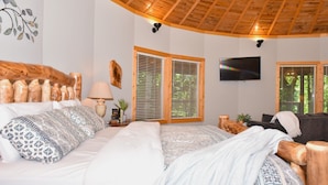 A bedroom with a wooden ceiling, two windows with blinds, a bed with patterned bedding, a TV on the wall, and a small table with a lamp and decorative items.