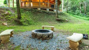 A wooden cabin with a stone fire pit and two stone benches in a forest setting.