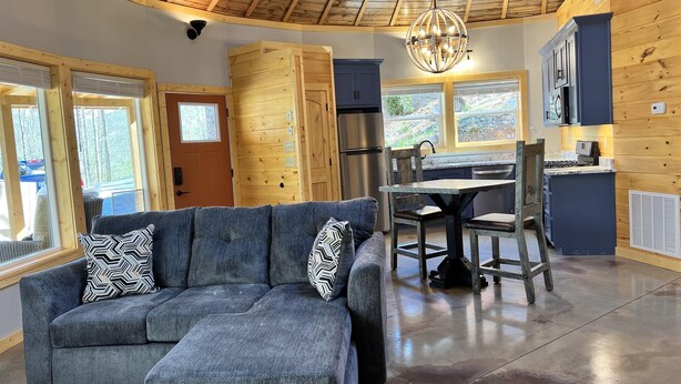 A modern cabin interior with a gray sectional sofa, wooden walls, a dining area, and a kitchen with dark cabinets.