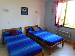 A room with two wooden beds, each with blue bedding and pillows, a small wall decoration, a window with pink curtains, and a radiator.