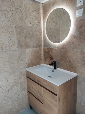 A bathroom with a white sink, wooden vanity, and a round mirror with a light above it.