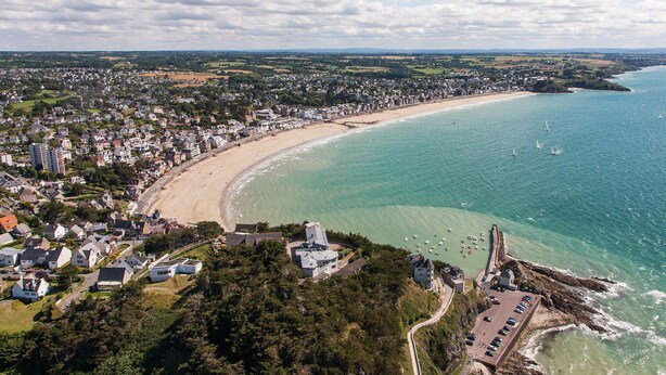 Vue aérienne d’une ville côtière avec une plage, des zones résidentielles et une côte rocheuse.