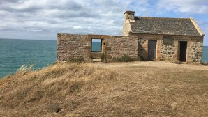 Un bâtiment en pierre avec un toit en tuiles, une petite fenêtre et une porte, situé sur une colline herbeuse surplombant une étendue d’eau.