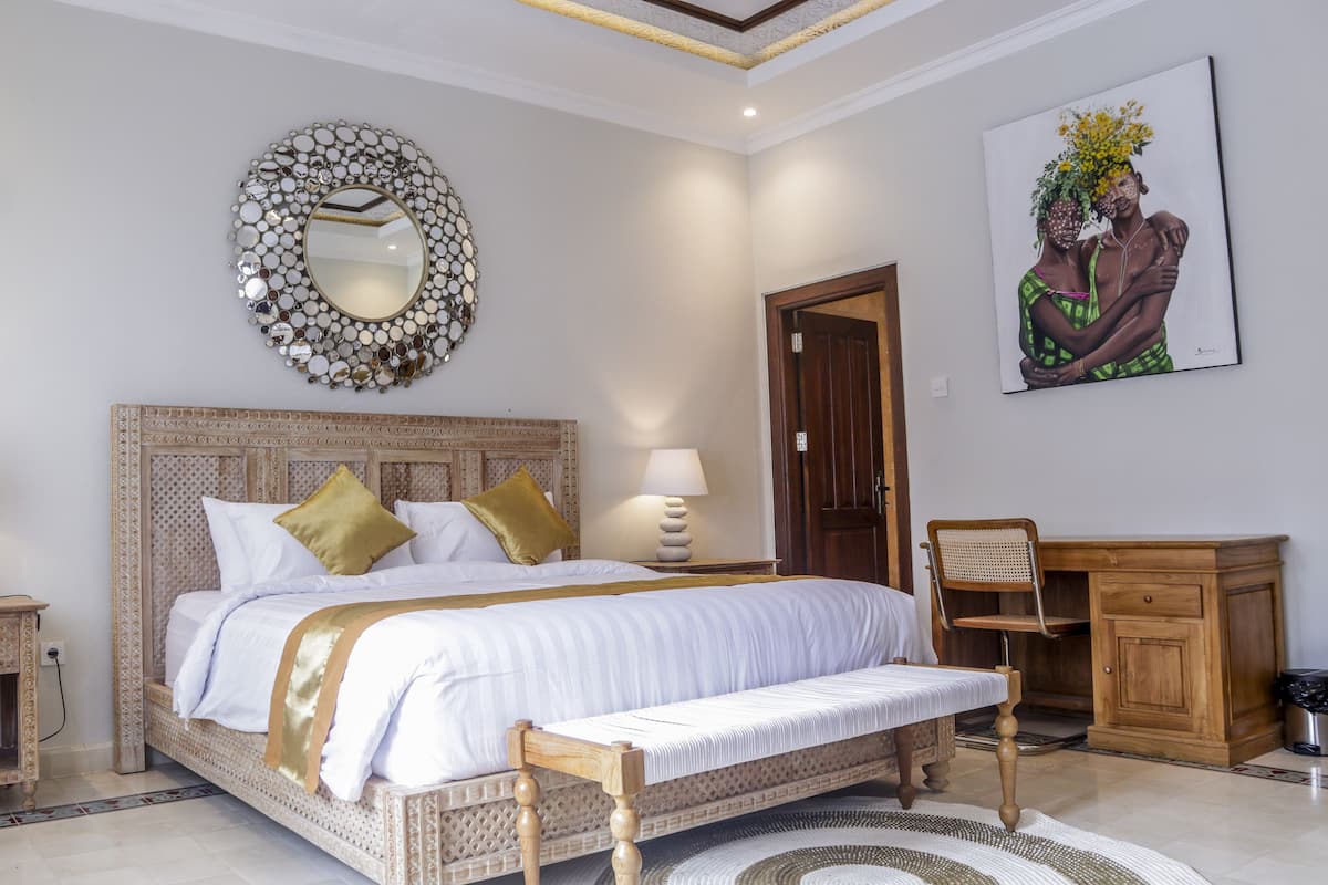 A bedroom with a bed, two bedside tables with lamps, a wooden bench, a decorative mirror, a wooden door, and a painting on the wall.