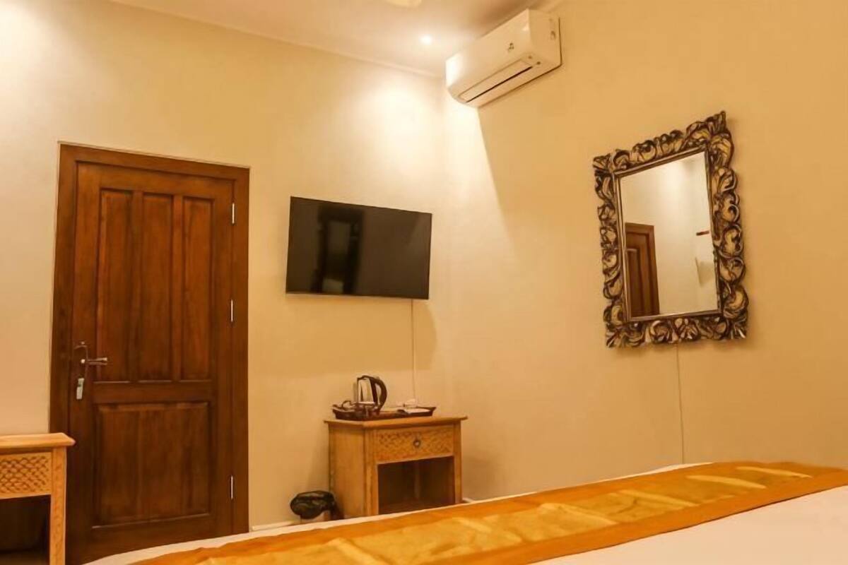 Quarto de hotel com porta de madeira, televisão, espelho e uma pequena mesa com chaleira.