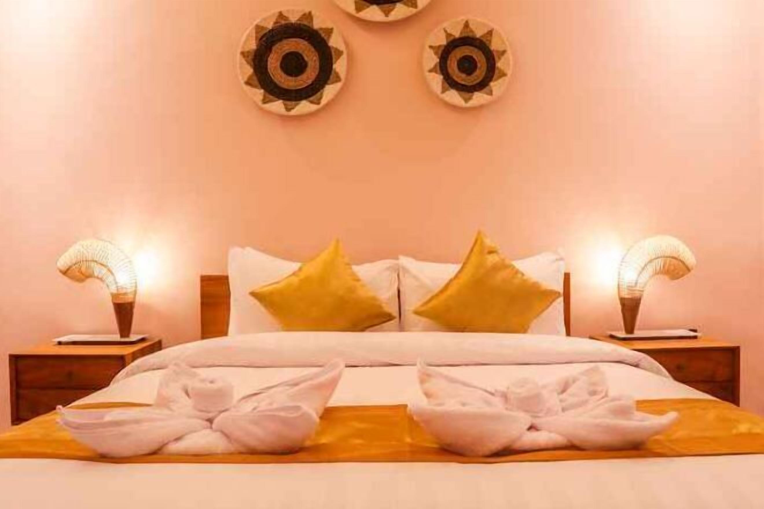 Cama bem arrumada com lençóis brancos e duas almofadas amarelas, com duas luminárias laterais sobre mesas de cabeceira de madeira.