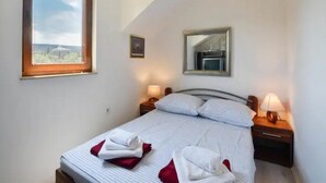Un lit bien fait, avec du linge de lit blanc et rouge, une lampe de chevet, une petite table de chevet, une fenêtre donnant sur un beau paysage et un miroir accroché au mur.