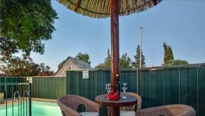Un espace aménagé au bord de la piscine, avec des chaises et une table en osier, un parasol en chaume et une clôture en arrière-plan.