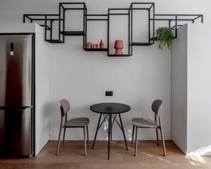 Una cocina moderna con refrigerador empotrado, una mesa de comedor redonda con dos sillas y una estantería metálica negra con jarrones rojos y una planta en maceta.