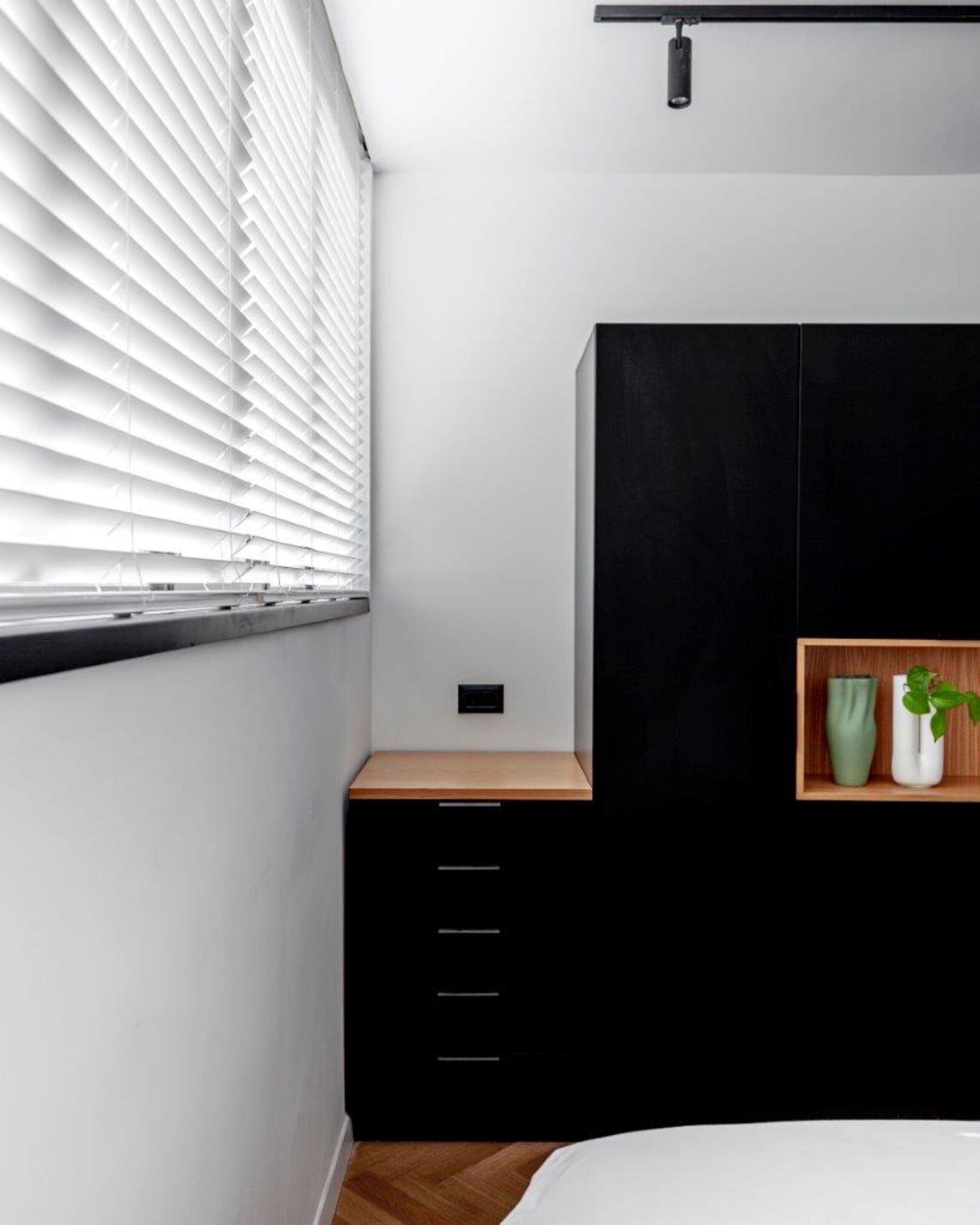 A bedroom with a bed, a black wardrobe, a wooden nightstand, a shelf with two vases, and a window with blinds.