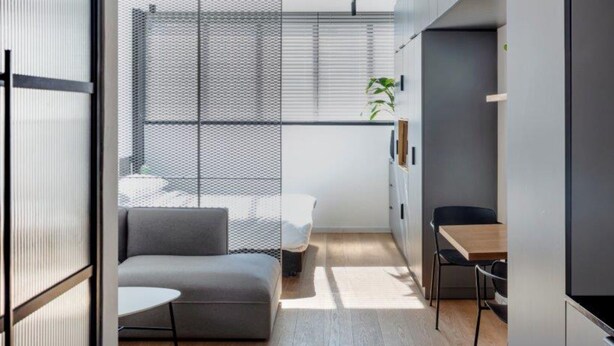 A modern, minimalist bedroom with a large bed, a small seating area, and a window with blinds. - DeBlox living - Ben Avigdor (Tel Aviv)