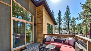 Una terraza de madera con un ventanal que da a una habitación, un sofá de mimbre con cojines y una mesa de vidrio.