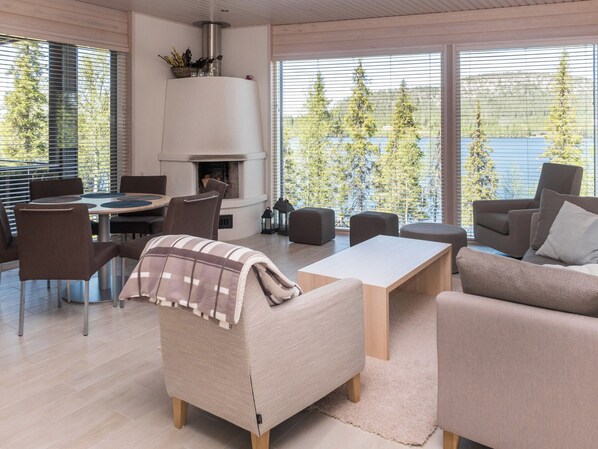 Un salon moderne doté d’une cheminée, de sièges confortables et de grandes fenêtres offrant une vue sur les arbres et un plan d’eau.