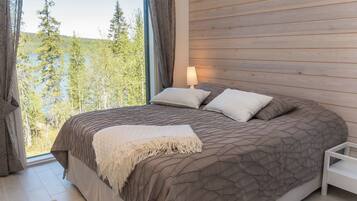 A bedroom with a large bed, a nightstand, a lamp, and a view of trees and a lake.