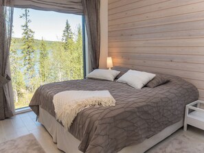 A bedroom with a large bed, a nightstand, a lamp, and a view of trees and a lake. - Holy lake rantasauna by Interhome (Kuusamo)